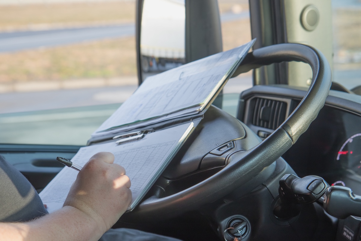 Truck driver in the cab of a big rig recording hours of service in his logbook.